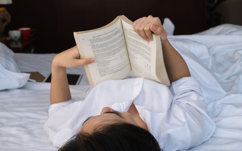 Femme allongée dans son lit sur le dos, lisant un livre.