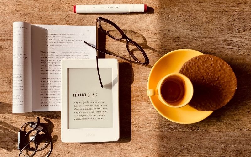 Photo prise de haut d'un livre papier, d'un livre numérique sur tablette, d'une paire de lunette, d'un feutre et d'une tasse de café.