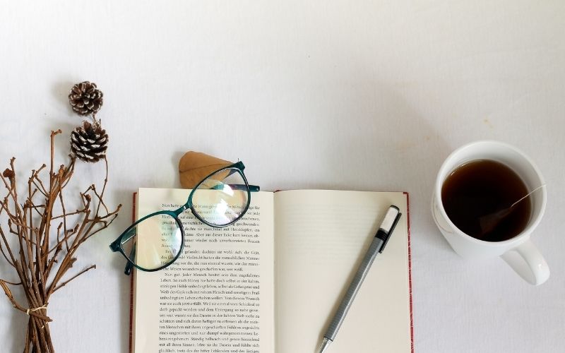 Photo d'un livre ouvert, posé sur une table blanche, avec une paire de lunette, une tasse de café à droite, des fleurs séchées à gauche, et un stylo sur le livre.